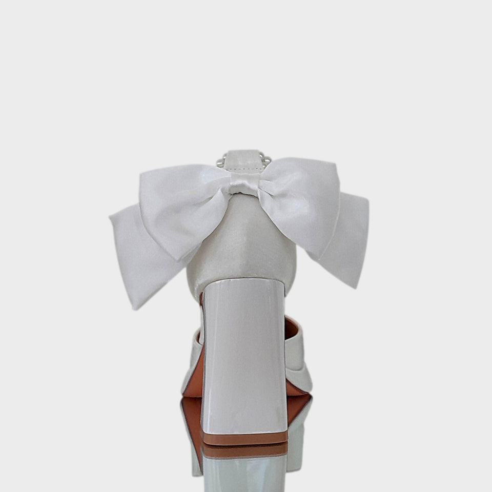 Vista de talón y taco de las plataformas Caroline en raso de color off white, con doble pulsera de perlas, gran moño de raso off white en el talón, y taco bloque en acrílico al tono de 9 cm