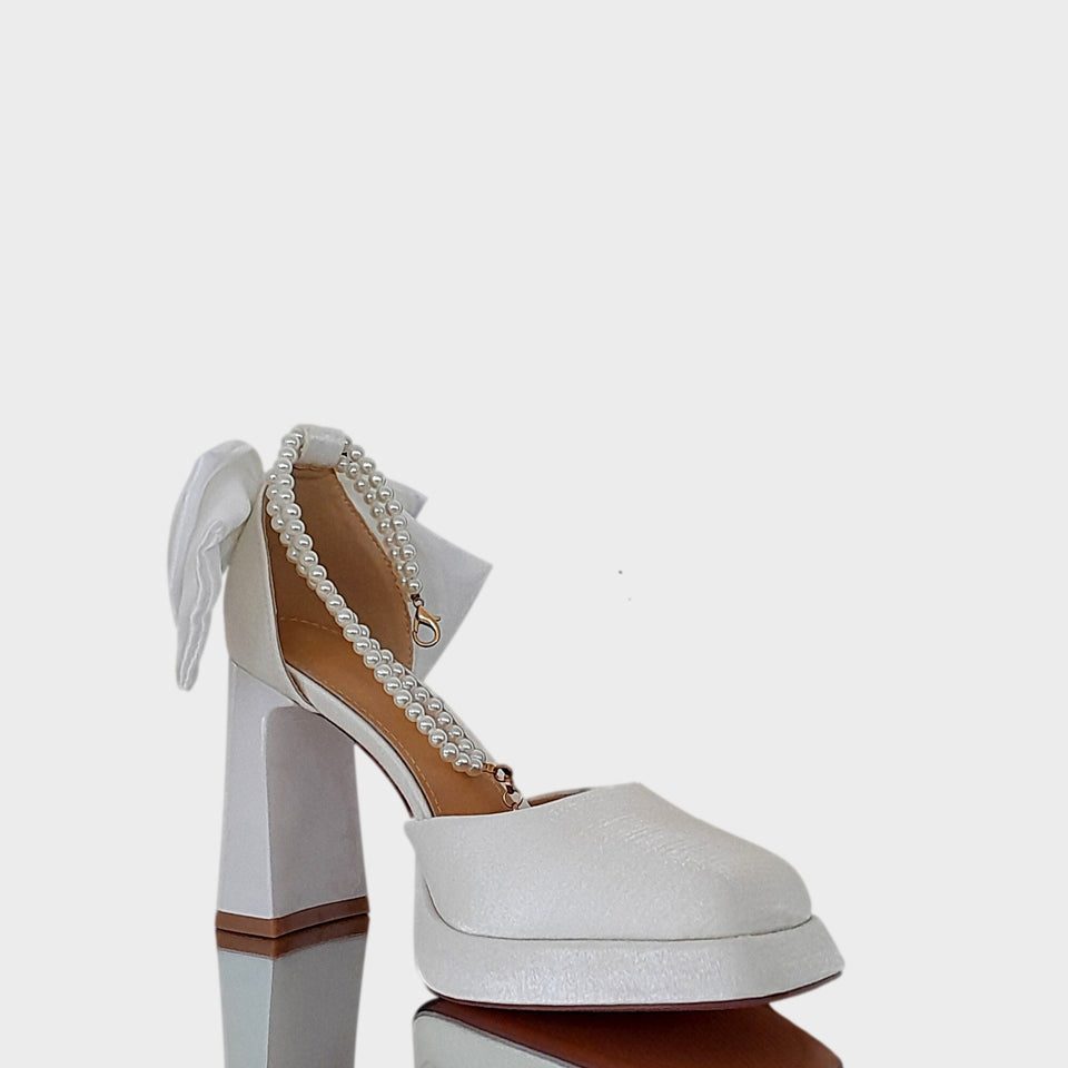 Vista lateral de las plataformas Caroline en raso de color off white, con doble pulsera de perlas, gran moño de raso off white en el talón, y taco bloque en acrílico al tono de 9 cm