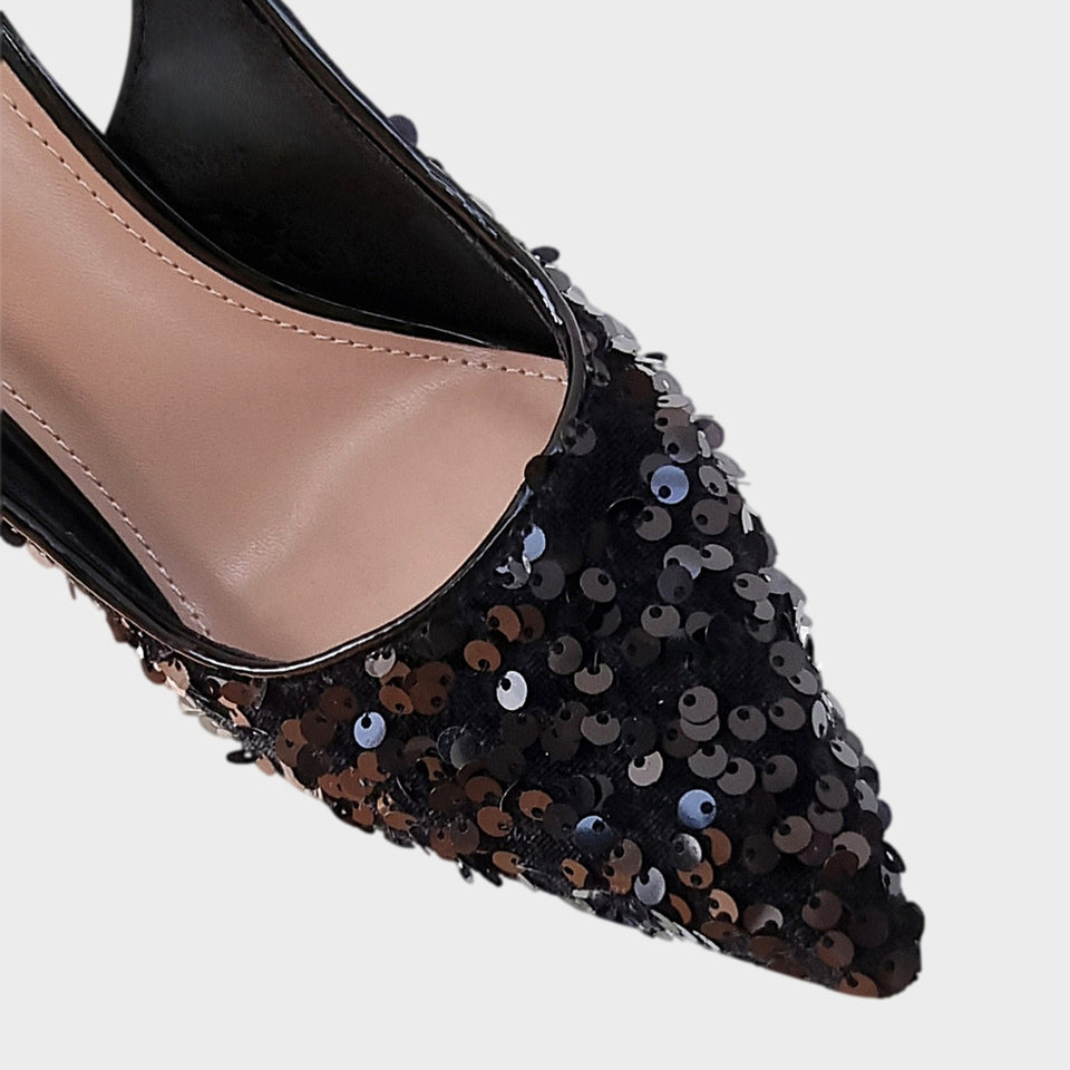 Vista en detalle de la capellada de elegantes stilettos de fiesta en lentejuelas de color negro, con talón descubierto y finísimo taco acrílico al tono de 10 cm